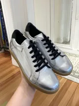 Zapatillas plateadas de cuero vacuno, con cordones negros, suela marrón y detalles en negro en el talón.