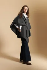 Poncho de tejido suave color beige con diseño envolvente y terminaciones con costuras decorativas en contraste color negro. Presenta un detalle de lazo frontal integrado.