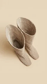 Botas de caña alta desmontables de gamuza color beige, con taco cuadrado de 6 cm y punta semi cuadrada. Presentan un diseño con costura central y detalle de estribo con hebilla forrada en el tobillo.