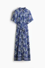 Vestido midi camisero de color azul con estampado ornamental en tonos blancos y azules. Cuenta con mangas cortas raglán, botonadura frontal completa y cinturón de la misma tela para atar en la cintura.