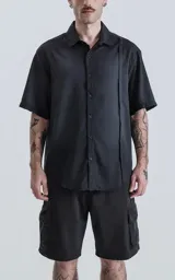 Camisa de manga corta color negro con diseño minimalista, cuello clásico y cierre frontal de botones. Presenta una costura vertical decorativa en el lado izquierdo del frente.