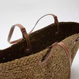 Bolso tipo capazo color kaki, confeccionado en tejido trenzado efecto rafia, con doble asa larga de hombro en cuero sintético marrón claro con costuras blancas visibles.