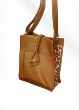 Bolso tipo matera o tote bag, color camel, con paneles laterales de estampado animal print (jirafa). Presenta un bolsillo frontal exterior y tiras de cuero trenzadas como detalle decorativo en la parte superior, además de correas de hombro ajustables.