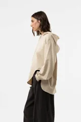 Buzo de punto oversize en color beige, con capucha, cordones largos ajustables y mangas largas amplias con puños elásticos. Presenta diseño asimétrico, más largo en la parte posterior.