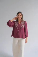 Blusa color bordó con bordados florales en tono crema en la parte superior frontal. Presenta cuello redondo con pequeña abertura, cierre frontal con botones y mangas largas acampanadas con puños ajustados.