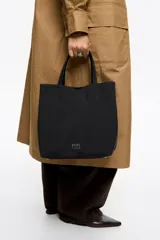 Bolso bandolera Bimba y Lola modelo Chihuahua de tamaño grande, color negro, con doble asa de mano y asa regulable de tejido. Cuenta con detalle de parche de cuero en un lateral, logo impreso en la parte inferior frontal y herrajes metálicos.