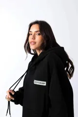 Hoodie negro de frisa con capucha y bolsillo canguro. Presenta un aplique rectangular en el pecho con texto en verde lima.
