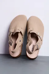 Sandalias estilo zueco color beige con tira trasera ajustable, hebillas metálicas plateadas, suela de corcho y base de goma negra.