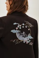 Saco sastre de color negro con corte clásico y solapas. Presenta un bordado artístico en la espalda con motivos de montañas, nubes y flores en tonos suaves.