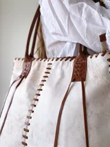 Cartera tipo tote de cuero vacuno con acabado texturizado en tono blanco hueso. Presenta detalles de costuras artesanales con tientos de cuero marrón en los bordes y correas de hombro a juego.