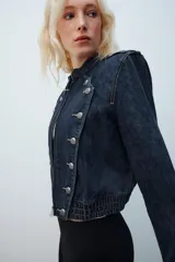 Chaqueta de denim de corte corto, color azul oscuro, con cuello redondo, hombreras y cierre frontal con cremallera. Presenta diseño de inspiración militar con doble botonadura metálica decorativa en el frente, trabillas con botón en los hombros y bajo elástico.