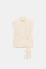 Camisa color crema de la colección Zara Woman, confeccionada en algodón 100%. Presenta cuello subido, manga sisa y un detalle de tejido tipo fajín asimétrico con un nudo en el lateral. El cierre frontal es con botones ocultos.