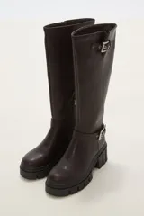 Bota de caña alta color negro, confeccionada en eco cuero, con suela track de goma y detalle de hebilla metálica en el tobillo y en la parte superior de la caña.