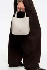 Bolso de mano de tamaño pequeño con estructura rígida, confeccionado en piel reconstituida color gris claro. Cuenta con doble asa superior, correa ajustable con logo de la marca y logo metálico en la parte frontal.