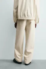 Pantalón chino de algodón color beige, de corte relajado (relaxed fit) y tiro medio. Presenta bolsillos delanteros y bolsillos traseros de vivo con cierre de cremallera y botón.