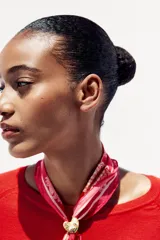 Pañuelo o bandana de seda y algodón, color fucsia intenso, con estampado floral y paisley en tonos beige/crema. Se anuda al cuello con un broche metálico en forma de corazón dorado y verde.