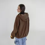 Canguro color marrón con capucha, cierre frontal parcial, bolsillo canguro y cordón ajustable en el dobladillo. Tiene un estampado blanco en el pecho con el texto "PULAU DESIGN DEPT." y un parche amarillo en la manga izquierda.