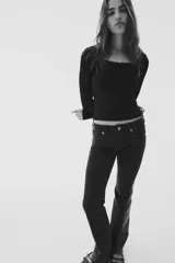 Remera negra de manga larga, corte ajustado y corto (cropped), confeccionada en tejido de punto acanalado. Presenta un escote redondo ribeteado con encaje y un pequeño lazo de satén en el centro del escote.