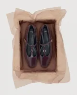 Zapato plano tipo Mary Jane, color bordó, de punta fina y tira en forma de T con hebilla metálica dorada ajustable.