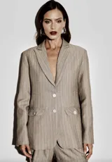 Blazer beige a rayas verticales, de corte recto y largo mediano, con solapa clásica, cierre de dos botones frontales, bolsillos con solapa y botones decorativos.