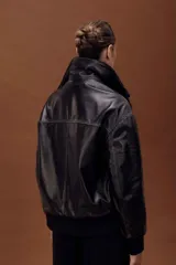 Chaqueta de cuero estilo bomber en color negro, con cuello alto, cierre frontal con cremallera y solapa con botones a presión. Presenta puños y cintura de tejido elástico acanalado, bolsillos laterales y un diseño estructurado.