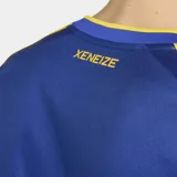 Camiseta de futbol Adidas de la primera equipacion de Boca Juniors, de color azul con la caracteristica franja horizontal amarilla en el pecho. Presenta cuello en V, mangas cortas con las tres tiras de Adidas en los hombros y el logo de la marca y el escudo del club bordados en el frente.