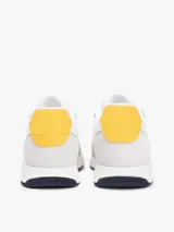 Zapatillas de estilo runner de Tommy Hilfiger, confeccionadas en una mezcla de cuero y textil en color blanco con detalles en gris claro y un panel amarillo en el talón. Presentan cordones blancos y suela de goma con base oscura.