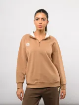Buzo de felpa color tostado con cuello alto y medio cierre frontal. Presenta un diseño de corte holgado con hombros caídos, puños y ruedo acanalados, y el logo de la marca bordado en el pecho.