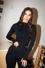 Campera deportiva entallada de color negro, con cierre frontal completo y cuello alto. Confeccionada en tejido mate de alta elasticidad, cuenta con mangas largas con aberturas para los pulgares.
