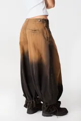 Jean de corte wide leg con efecto degradado en tonos marrón y negro. Presenta costuras verticales frontales y un diseño de puño ajustable en el bajo de la pierna.
