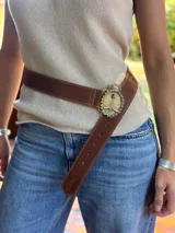 Cinturón de cuero marrón grueso con pespuntes, ajustado a la cintura. La hebilla es redonda, de metal dorado o bronce, con el Escudo Nacional Argentino en relieve rodeado por una corona de laureles y un sol en la parte superior.