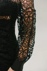 Vestido corto de encaje negro con diseño calado, escote en V, mangas largas con puños y falda con volados. Presenta un corte ajustado con detalle fruncido en la cintura y cierre invisible.