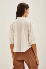 Camisa de algodón color celeste con cuello alto con volados, mangas tres cuartos abullonadas y cierre frontal con botones. Presenta un diseño con dos franjas verticales de bordado intrincado en color negro en el frente.