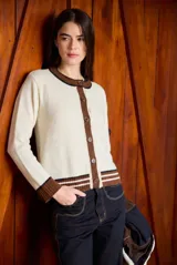 Cardigan clásico de largo medio color crudo, de manga larga, con cuello redondo, puños y ribetes en marrón con rayas blancas, y cierre frontal de botones marrones.