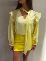 Camisa de tela semitransparente color amarillo claro, con mangas largas abullonadas, puños elásticos y detalle de volados en los hombros. Presenta un cuello con lazo integrado que cae sobre el frente.