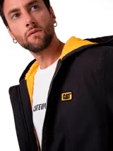 Chaqueta negra de lona con capucha, cierre frontal completo y forro interior en color amarillo. Presenta el logo de la marca en un parche rectangular en el pecho.