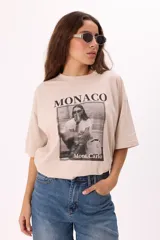 Remera color beige de corte amplio con mangas cortas y cuello redondo. Estampado frontal con la palabra "Monaco" en la parte superior y una fotografía en blanco y negro con la frase "Mont Carlo" en la parte inferior.