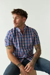 Camisa de manga corta con cuello abotonado, confeccionada en algodón con estampado de cuadros en tonos azules, rojos y blancos. Presenta un bolsillo frontal en el pecho con logo bordado y cierre de botones.