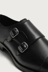 Zapato de vestir tipo monk de color negro, con detalle de doble hebilla metálica plateada en el empeine, puntera redondeada y suela con ligero tacón.