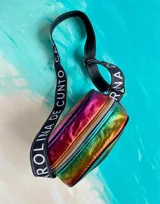 Bandolera Kaira de la línea Ottenta de Carolina De Cunto, confeccionada en material metalizado con acabado multicolor y efecto tornasolado. Cuenta con un bolsillo frontal con broche imantado, cierre superior y una correa larga y ajustable de tela negra con el nombre de la marca en letras blancas.