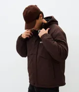 Campera tipo parka color marrón con capucha estructurada, cierre frontal y bolsillo vertical con cierre en el pecho. Presenta detalles de estampado utilitario en el frente y recortes en el cuerpo.