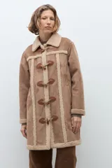 Abrigo de estilo duffle coat en simil gamuza color tostado, con interior y bordes de piel sintética al tono. Presenta cierre frontal mediante alamares de cuero sintético y madera, cuello solapa y mangas largas.