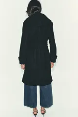 Abrigo de paño negro con capucha, cierre frontal cruzado con botones y cinturón de lazada en el mismo tejido. Presenta manga larga con trabillas en los puños y bolsillos laterales.