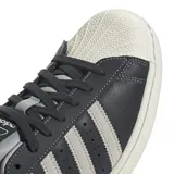 Championes adidas Superstar de diseño clásico con exterior de cuero sintético en color gris oscuro, icónica puntera de goma y suela de plataforma en tono crema. Presenta las tres bandas laterales en un tono gris más claro y el nombre del modelo en letras plateadas.