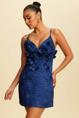 Vestido corto de tirantes finos en color azul profundo, confeccionado en tela satinada con textura arrugada y diseño de hojas en relieve. Presenta apliques florales tridimensionales en el escote y el frente, con un corte ajustado que realza la silueta.