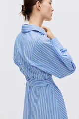 Vestido midi camisero de rayas azules y blancas, con cuello clásico, mangas largas, cierre frontal de botones y lazo ajustable en la cintura.