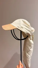 Gorro tipo pañuelo de lino color crudo con visera rígida en tono tostado. Presenta un bordado sutil con el número 1986 en el lateral y diseño envolvente con caída larga en la parte posterior.