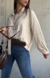 Camisa de corte holgado en color beige, con cuello clásico, cierre frontal de botones y mangas largas que presentan un delicado bordado tonal en los antebrazos.