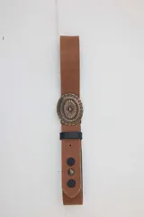 Cinturón de cuero color gris con efecto desgastado, hebilla metálica grande y redonda con diseño grabado, pasador marrón y tachas decorativas.