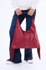 Cartera tipo hobo color beige, confeccionada en cuero sintético, con flecos laterales y detalle de tachas metálicas en el frente.